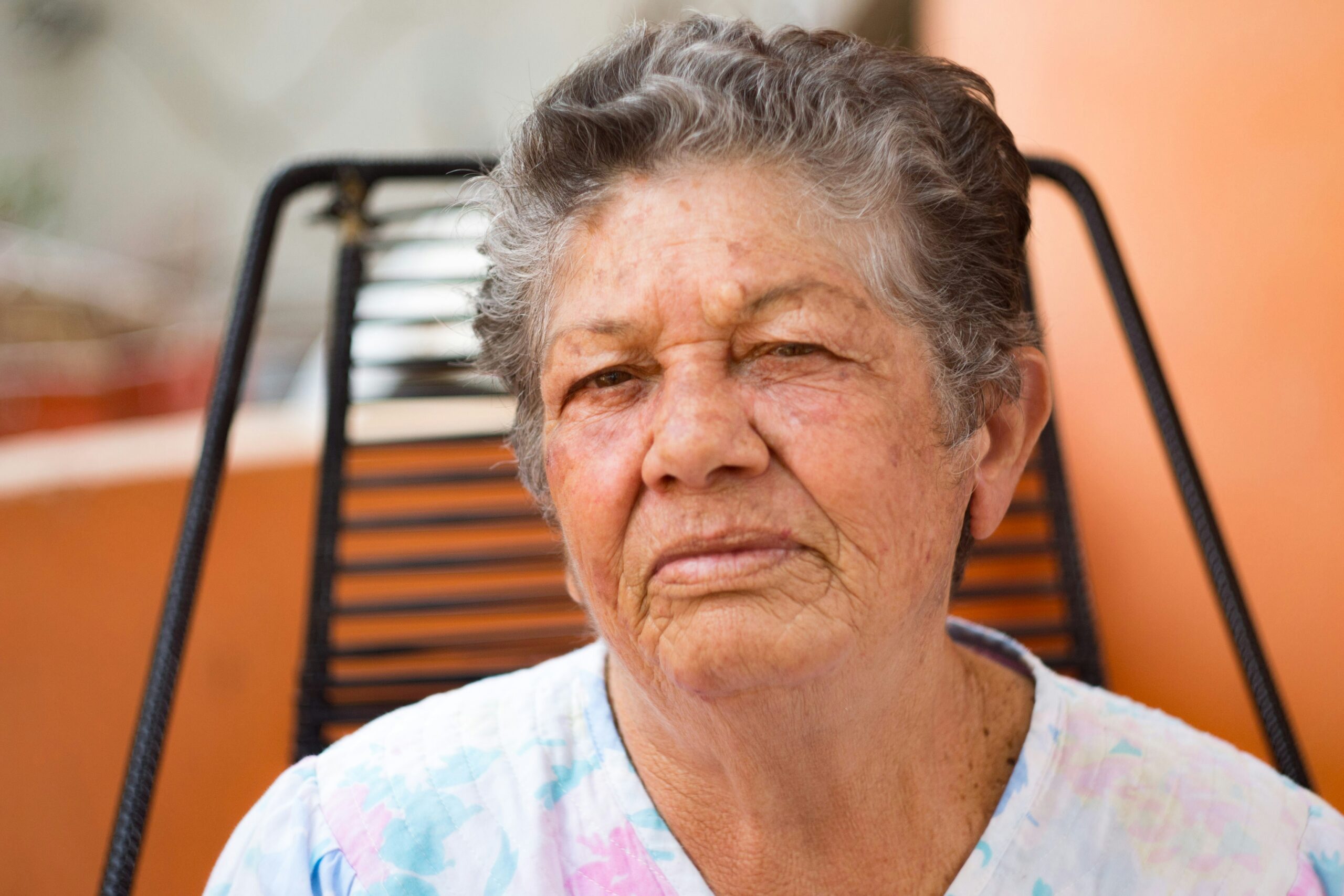 abuela | Ruiz Healy Times Close-up portrait of an elderly woman with gray hair, seated on a black metal chair against an orange background.