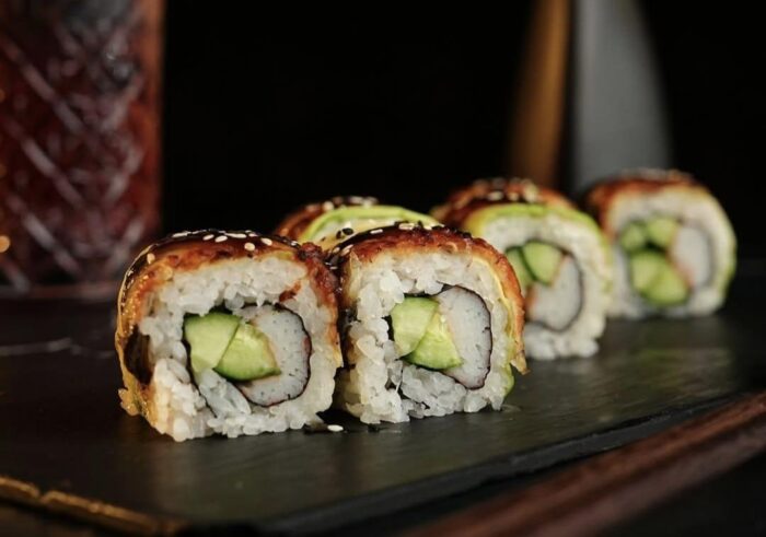 image | Ruiz Healy Times Sushi rolls with avocado filling on a slate plate, sesame seeds on top, close-up view in a dark setting.