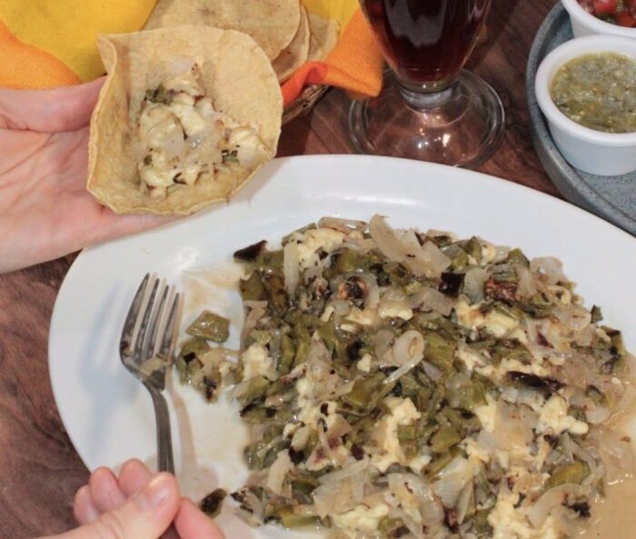 image | Ruiz Healy Times Hand holding a tortilla filled with a creamy chopped filling over a plate of sautéed green peppers and onions with melted cheese.