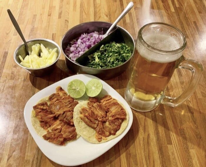 image | Ruiz Healy Times Two street-style pork tacos on a white plate with lime wedges, pineapple, onions and cilantro, beside a beer mug on a wooden table.