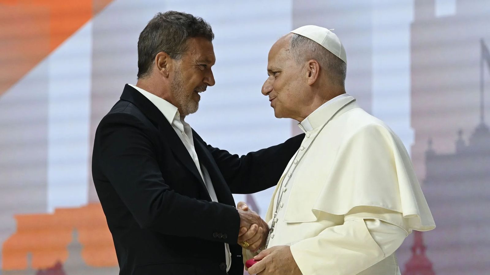 Two men in a handshake: a man in a dark suit shaking hands with Pope Francis in white papal robes, both smiling on stage.