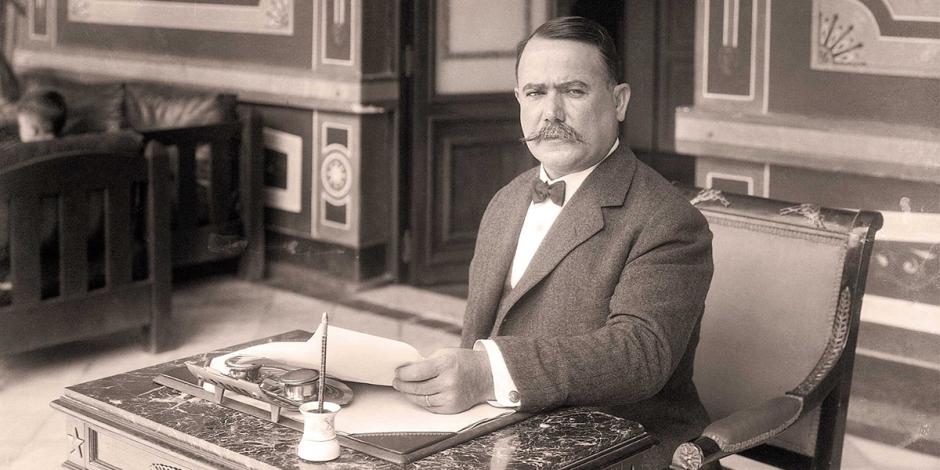 Historically dressed man with a mustache sits at a carved desk, pen in hand, in a formal office setting.