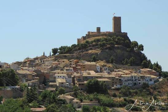 Medieval castle perched on a hill above a sunlit village with stone houses and greenery.