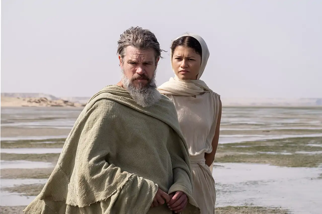 Bearded man in a beige cloak and a woman with a light headscarf stand on a barren salt-flat landscape under a pale sky.