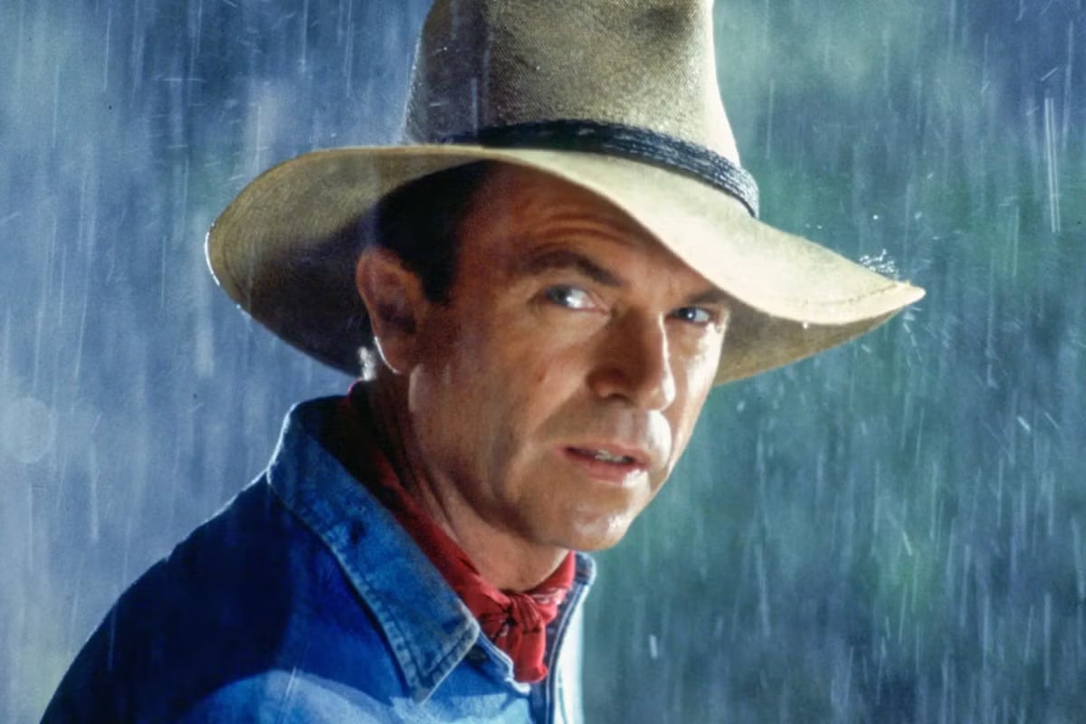 Man in a wide-brimmed hat and blue denim shirt stands in heavy rain, looking toward the camera.