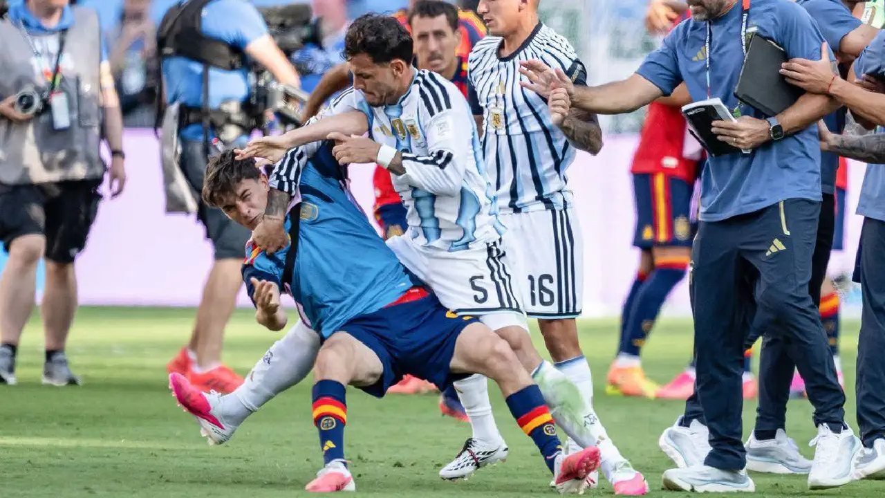 Soccer players collide as a blue jersey player falls while an opponent in striped white and blue reaches in for a tackle; staff and cameras on the sidelines