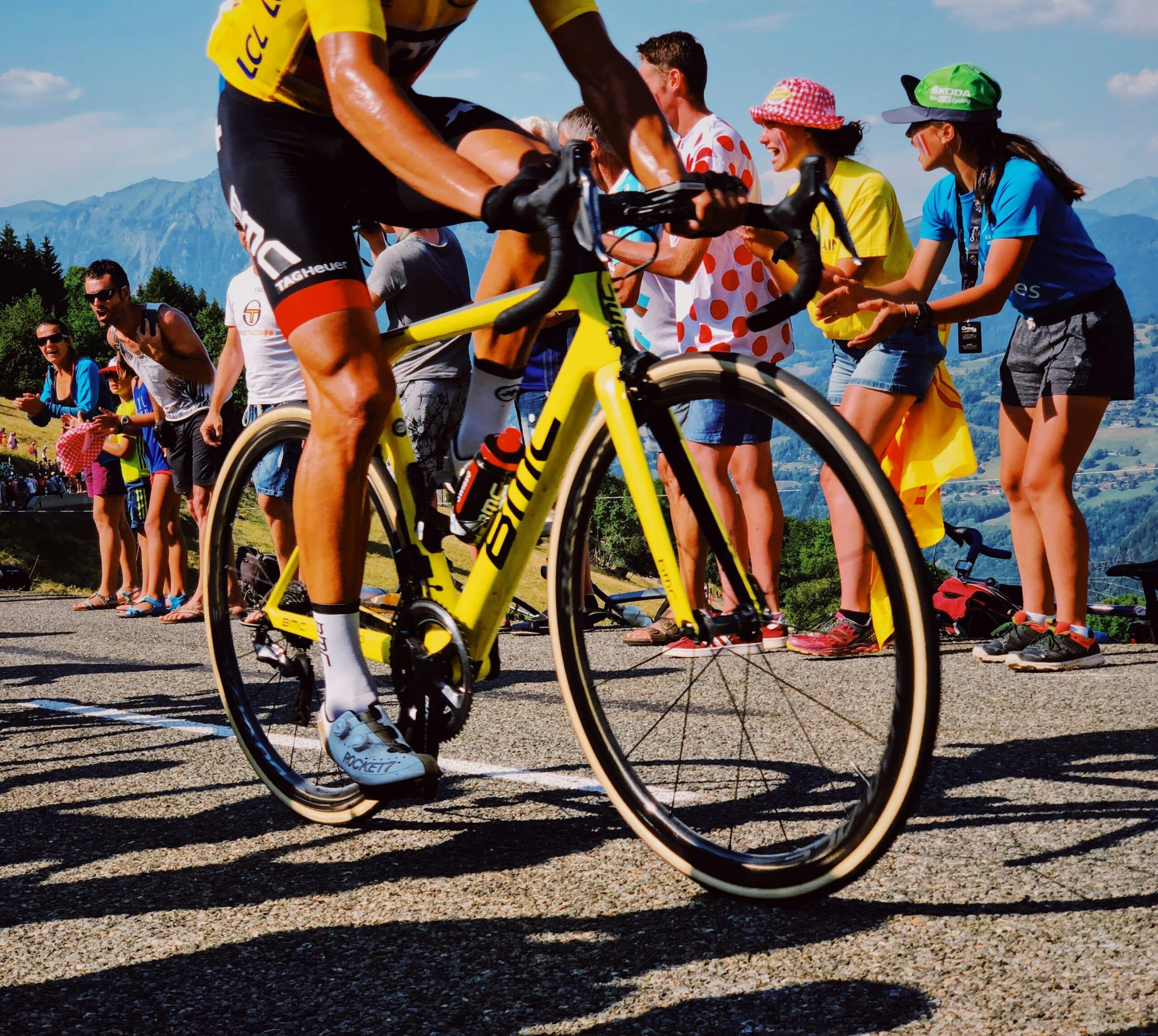 Close-up of a yellow racing bike wheel and cyclist's legs at the start of a race, with cheering spectators along a mountain road in the background.