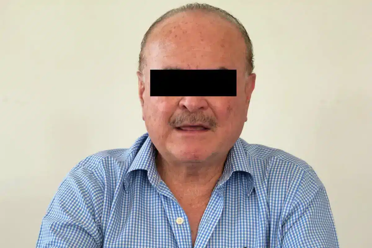 Adult man with a black bar over his eyes, wearing a blue checkered shirt.