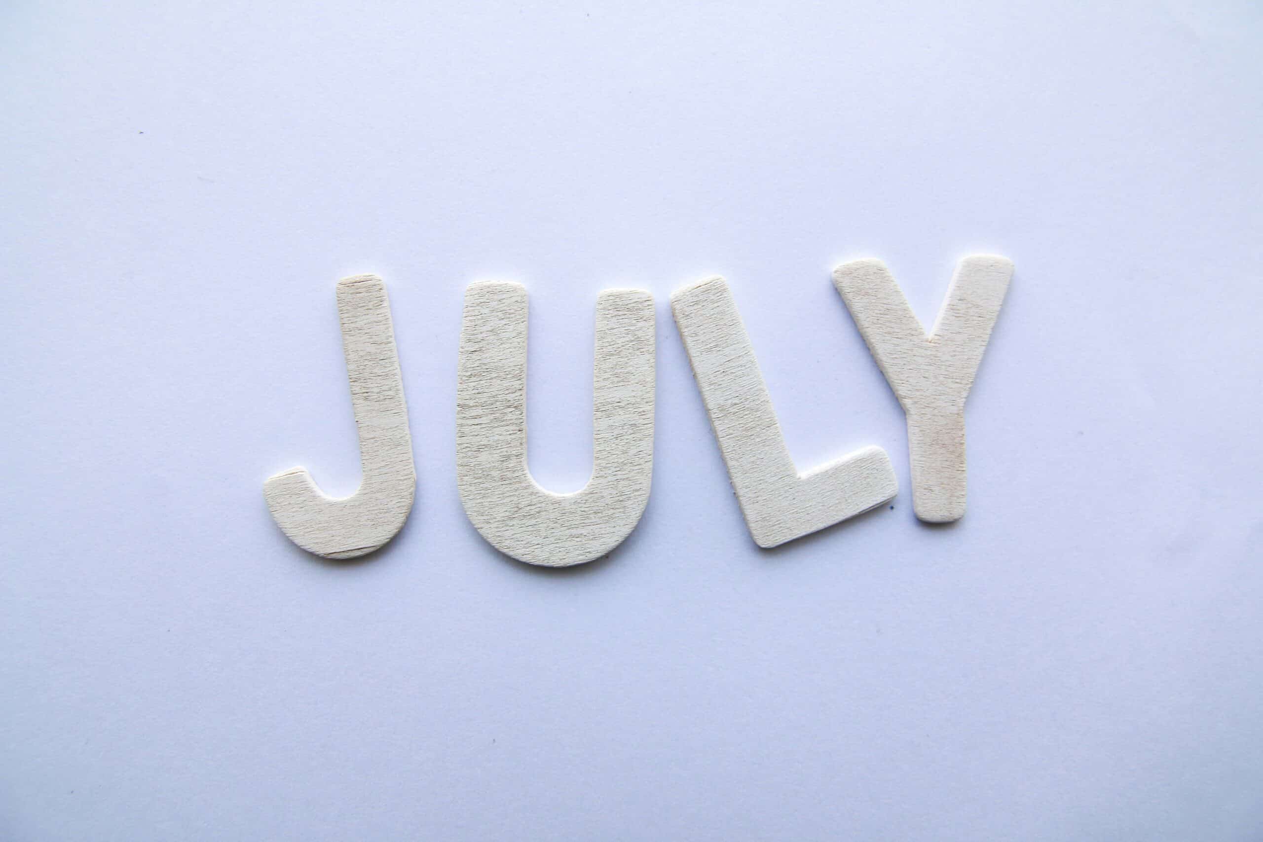 Wooden letters spelling 'JULY' arranged along a light purple background.