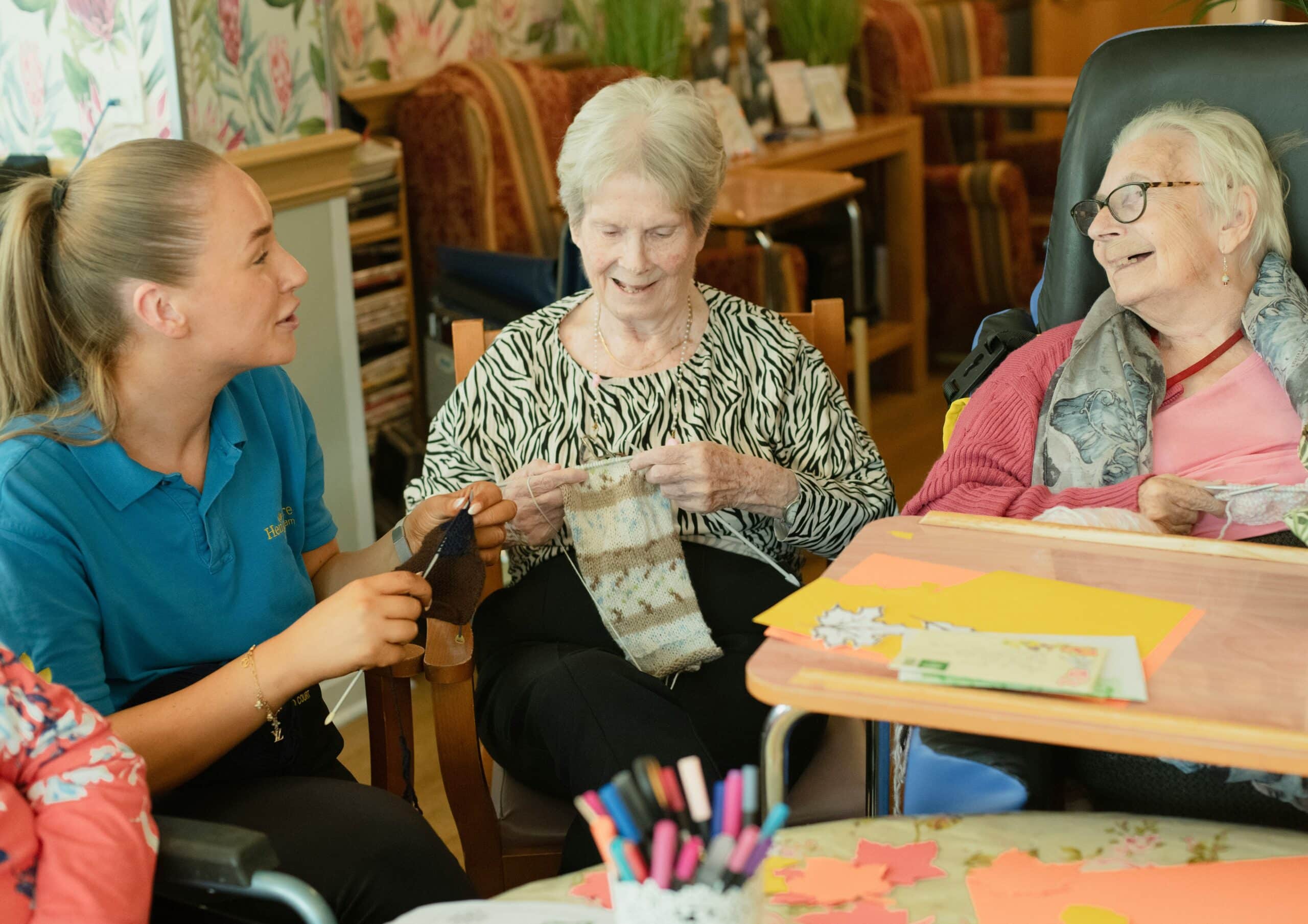 Caregiver speaks with two elderly women during a knitting activity at a craft table in a senior center.