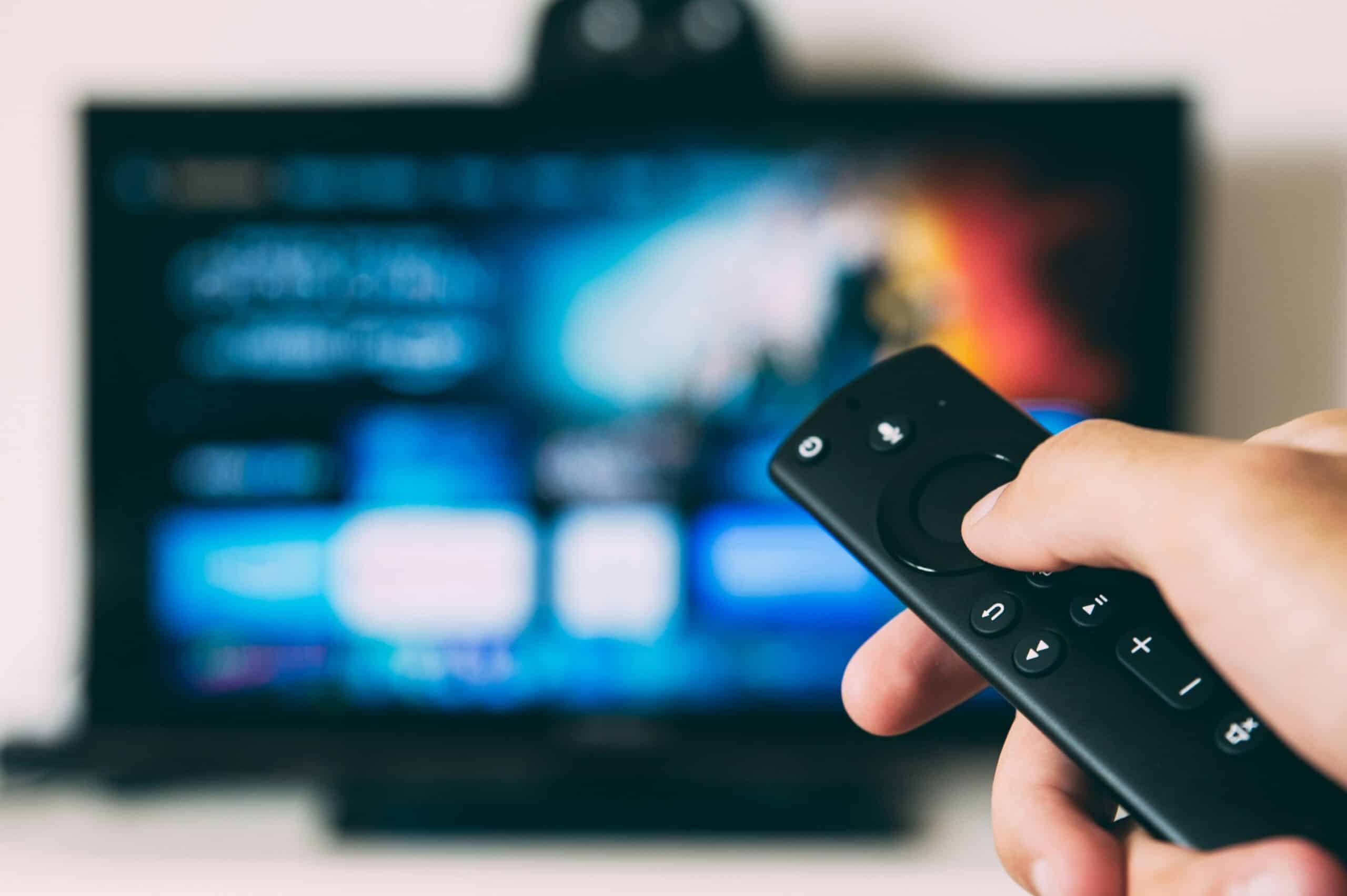 Hand holding a TV remote in focus with a blurred television screen in the background, suggesting changing channels or volume control.