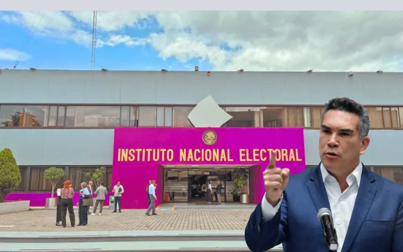 Man in a dark suit at a podium speaks into a microphone outside a bright pink INSTITUTO NACIONAL ELECTORAL building, with people gathered nearby