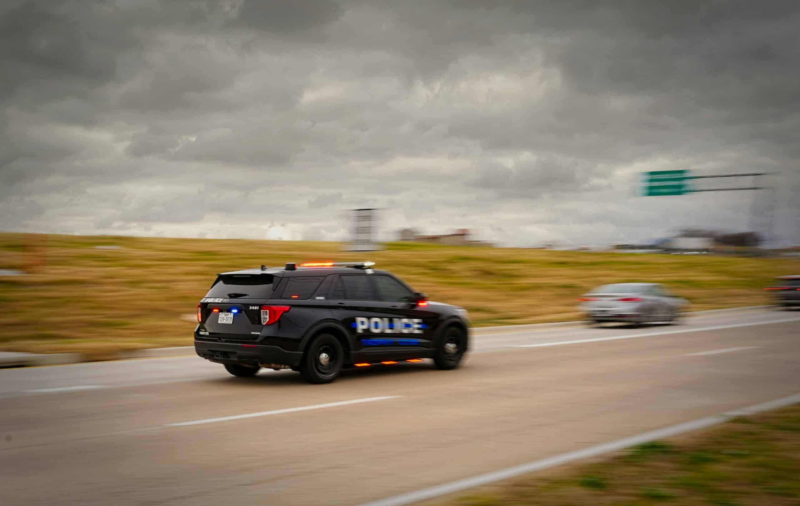Police SUV with flashing lights speeding along a highway as dark storm clouds loom overhead.