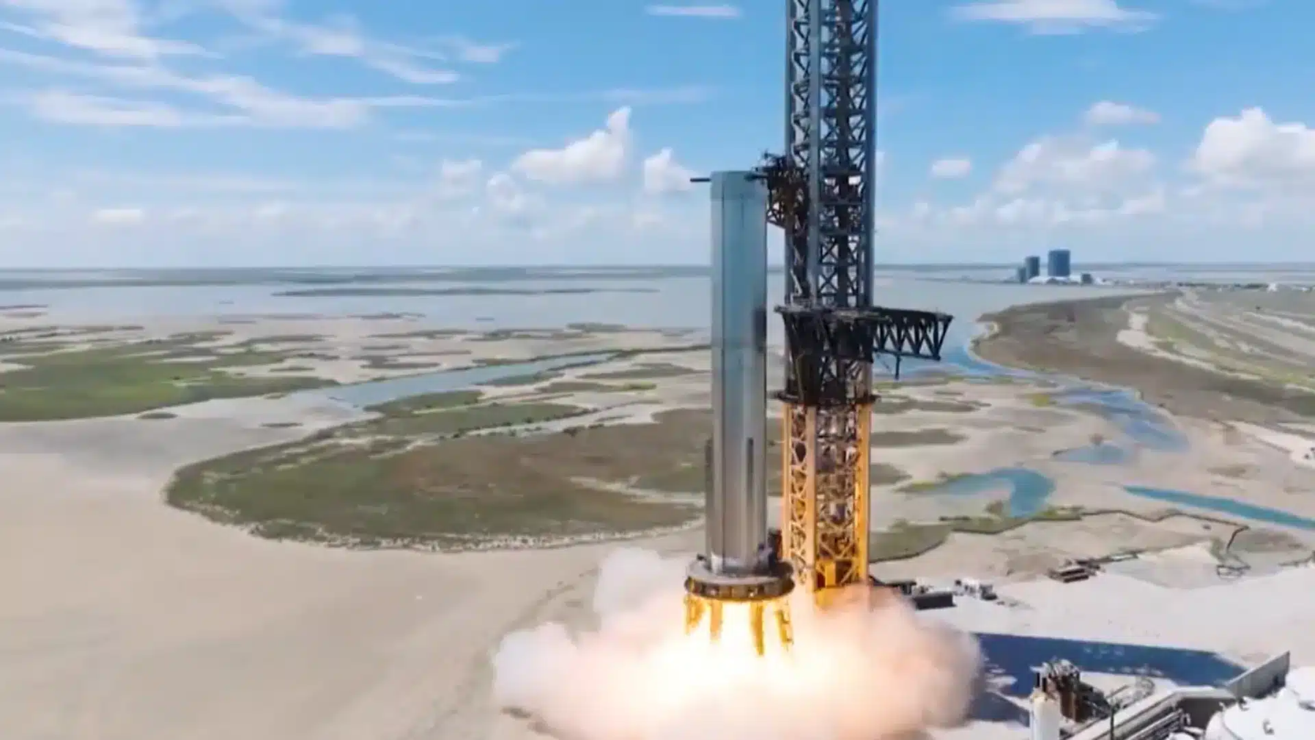 Rocket lifting off from a tall launch tower on a coastal marshland, with smoke plume at liftoff and blue sky above